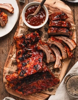 A rack of root beer ribs, partially sliced, on a wooden cutting board with an extra bowl of sauce.