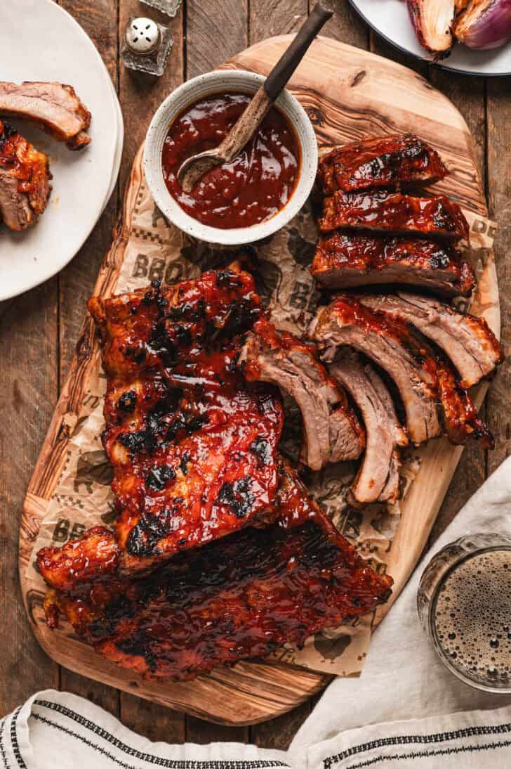 A rack of root beer ribs, partially sliced, on a wooden cutting board with an extra bowl of sauce.