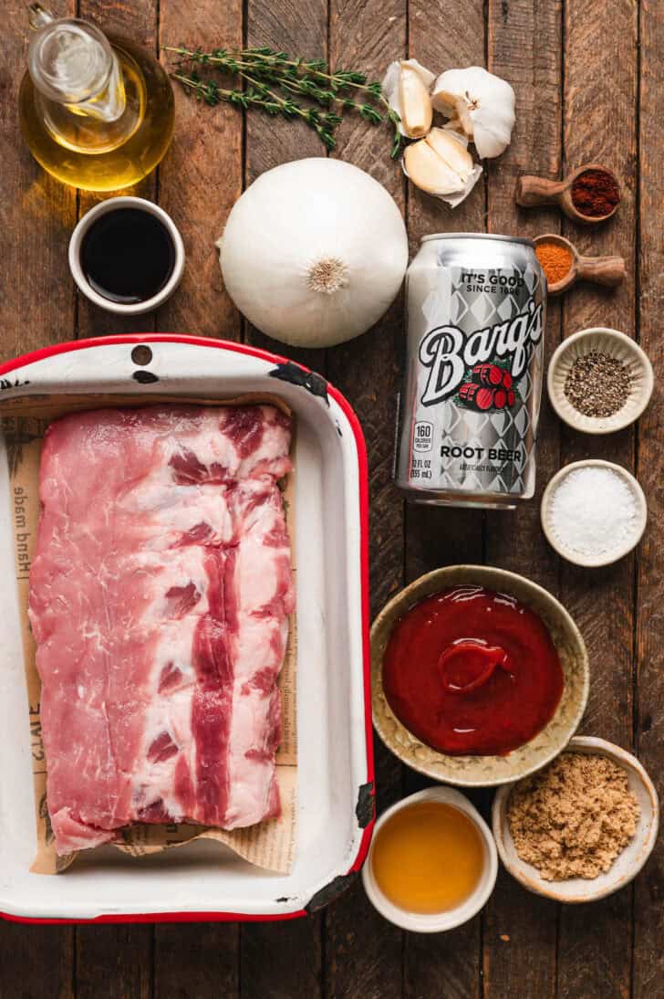 Ingredients for a pork rib marinade on a wooden surface including raw meat, ketchup, soda, onion, garlic, herbs, oil and spices.