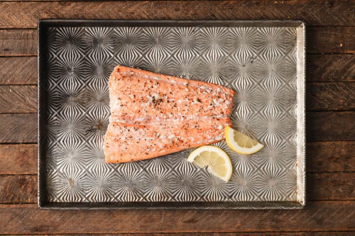 A piece of roasted orange-hued fish on a baking pan with lemon wedge