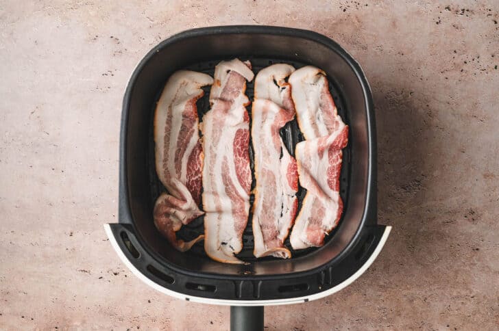 A photo demonstrating how to cook bacon in air fryer. Raw bacon is arranged in a single layer in air fryer basket.