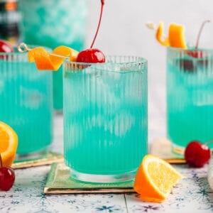 Three blue drinks garnished with orange peels and cherries.