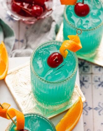 Three blue drinks garnished with orange peels and cherries.