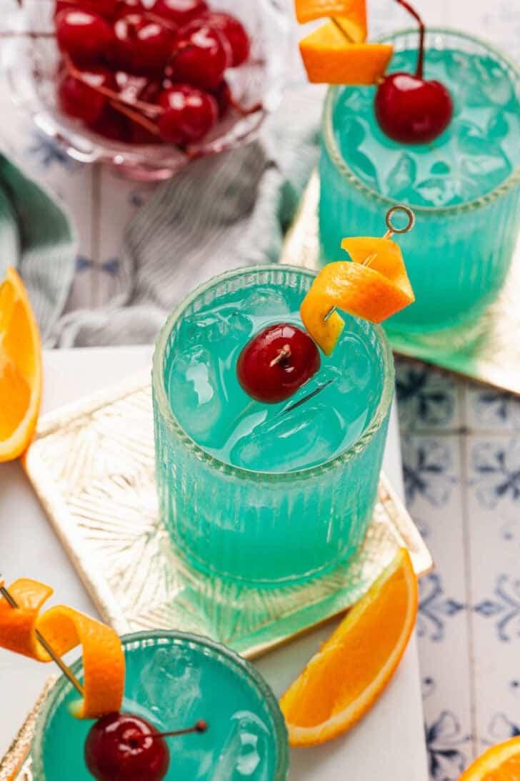 Three blue drinks garnished with orange peels and cherries.