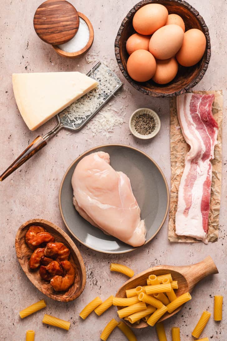 Ingredients on a light surface, including raw poultry, smoked shrimp, bacon, eggs, pasta, cheese and spices.
