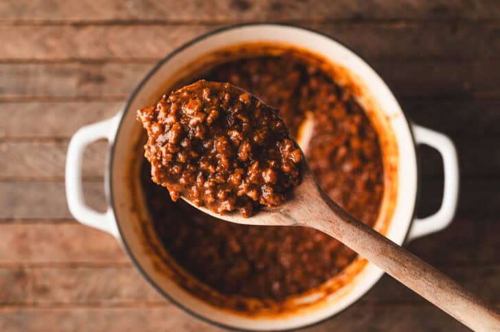 A spoon lifting a scoop of a chili recipe for hot dogs over a white pot.