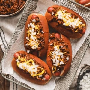 Four hot dogs in a basket covered in a hot dog chili recipe, cheese and onions.