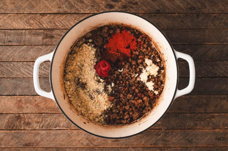 A white Dutch oven filled with cooked ground beef topped with tomato paste, breadcrumbs and spices.