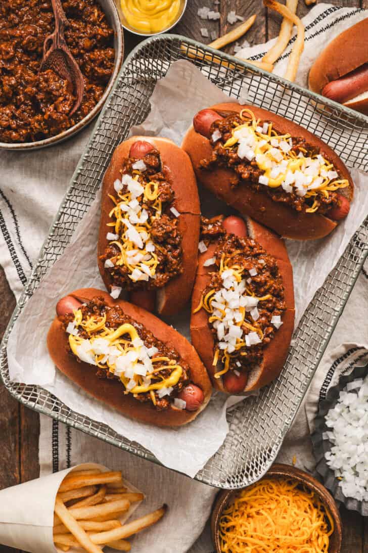 Four hot dogs in a basket covered in a hot dog chili recipe, cheese and onions.