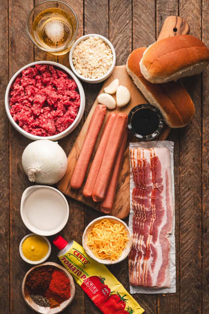 Ingredients on a wooden surface, including sausages, bacon, meat, spices, onions and beer.