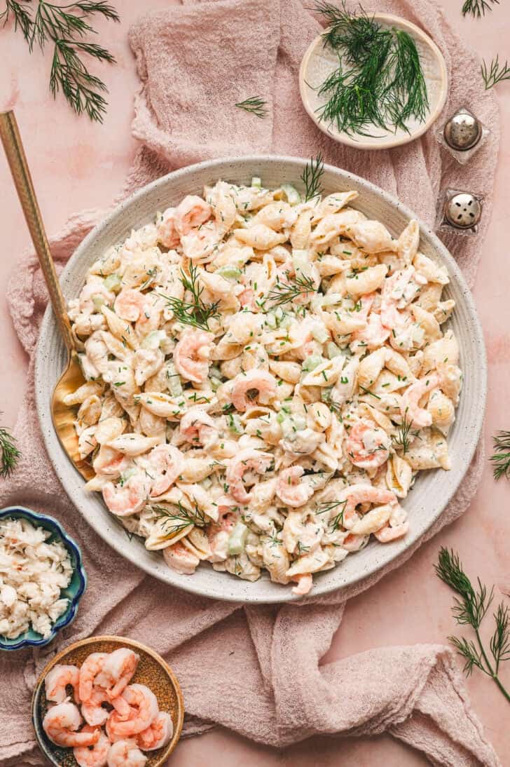 A bowl of seafood pasta salad on a pink surface with garnishes around it.