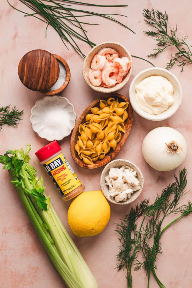 Ingredients on a pink background, including noodles, shrimp, crab, vegetables, mayonnaise, spices and herbs.