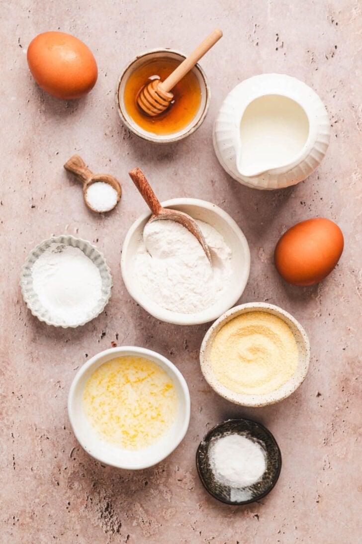 Ingredients on a texture surface, including flour, corn meal, leaveners, honey, eggs, butter and buttermilk.