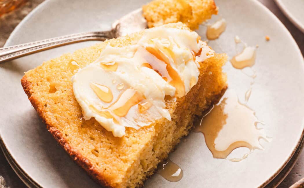 A slice of honey cornbread topped with butter and honey on a small white plate.