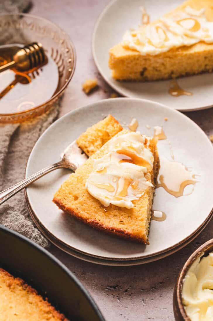 A slice of honey cornbread topped with butter and honey on a small white plate.