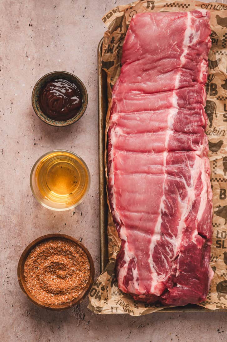 A raw slab of pork meat, with ramekins of barbecue sauce, apple juice and spice rib next to it.