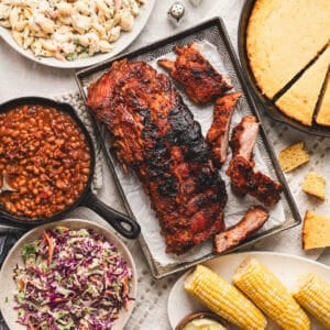 An overhead photo of baby back ribs surrounded by sides for ribs like corn, cornbread and cold salads.