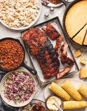An overhead photo of baby back ribs surrounded by sides for ribs like corn, cornbread and cold salads.