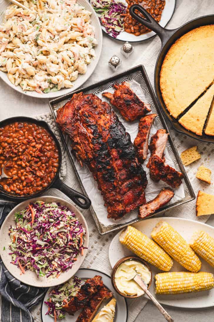 An overhead photo of baby back ribs surrounded by sides for ribs like corn, cornbread and cold salads.