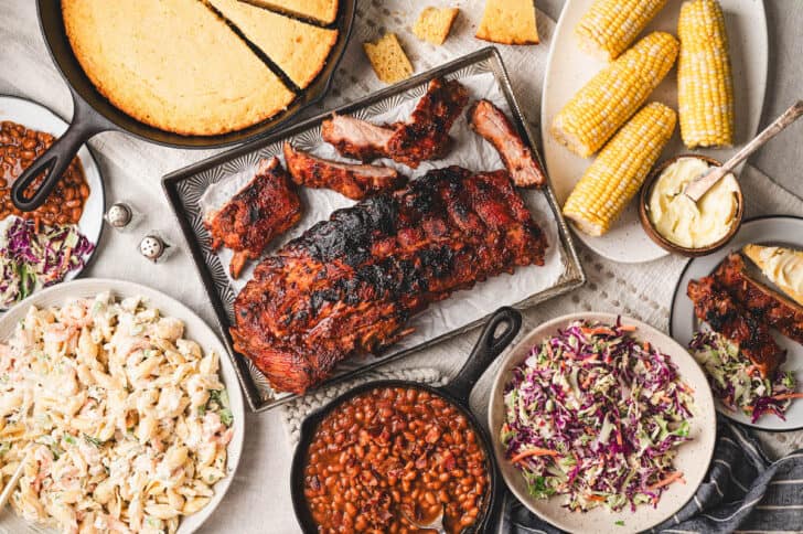 An overhead photo of baby back ribs surrounded by sides for ribs like corn, cornbread and cold salads.