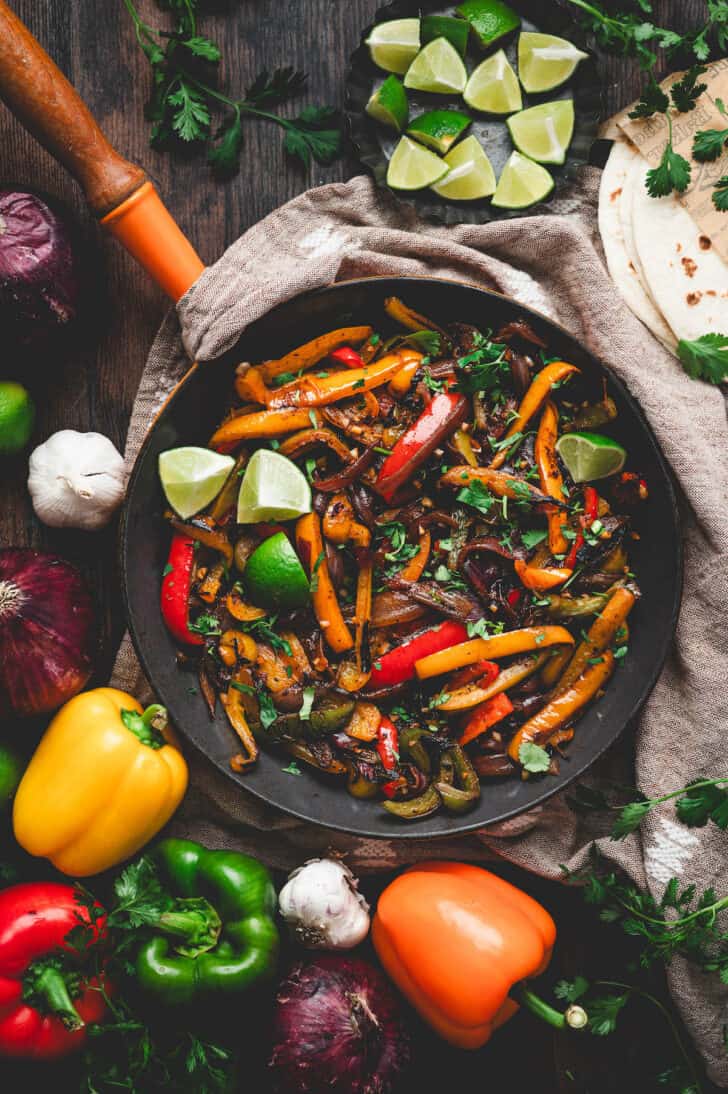 A skillet of fajita veggies surrounded by bell peppers and lime wedges.