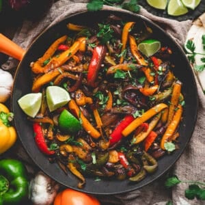 A skillet of fajita veggies surrounded by bell peppers and lime wedges.