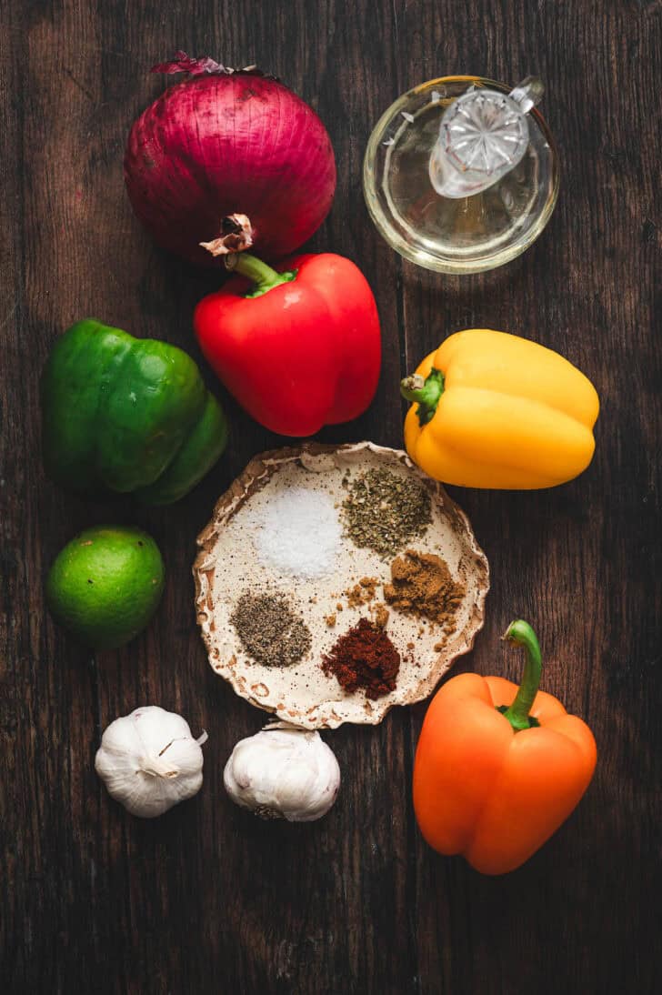 Bell peppers, onion, garlic, lime and spices on a dark tabletop.