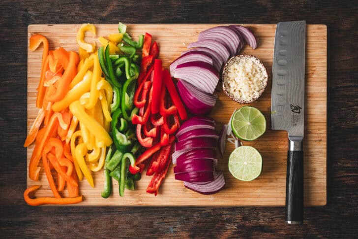 Sliced bell peppers and onions, chopped garlic and halved limes on a wooden cutting board.
