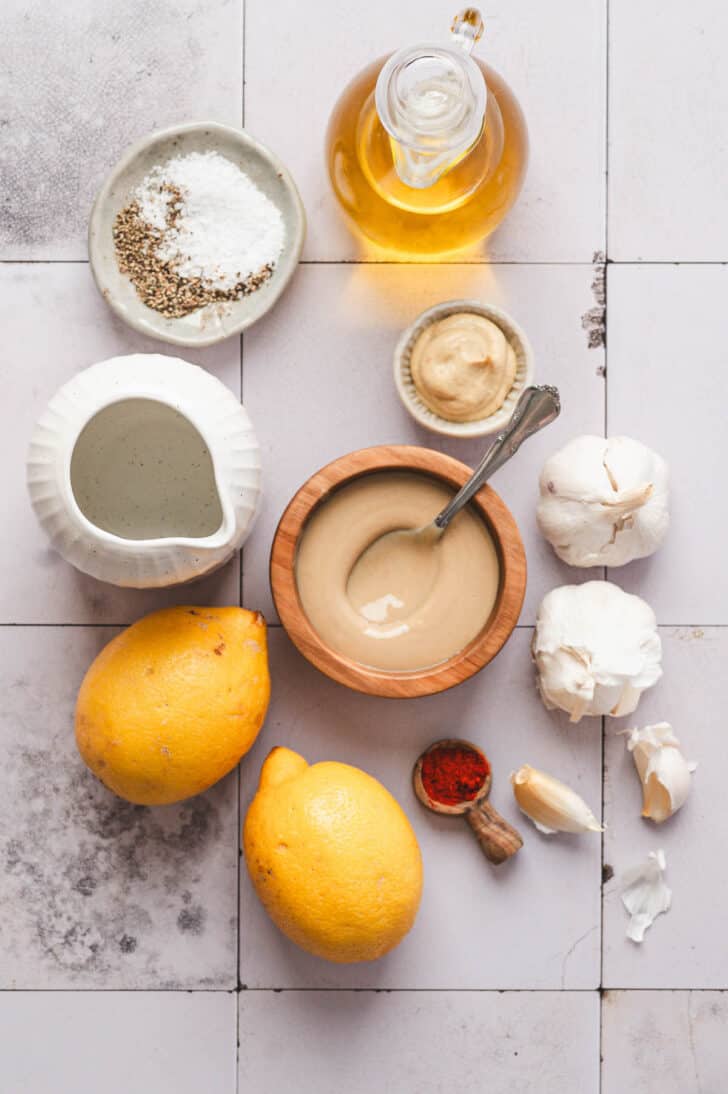 Ingredients on a light tiled surface, including sesame seed paste, lemons, garlic, oil and spices.