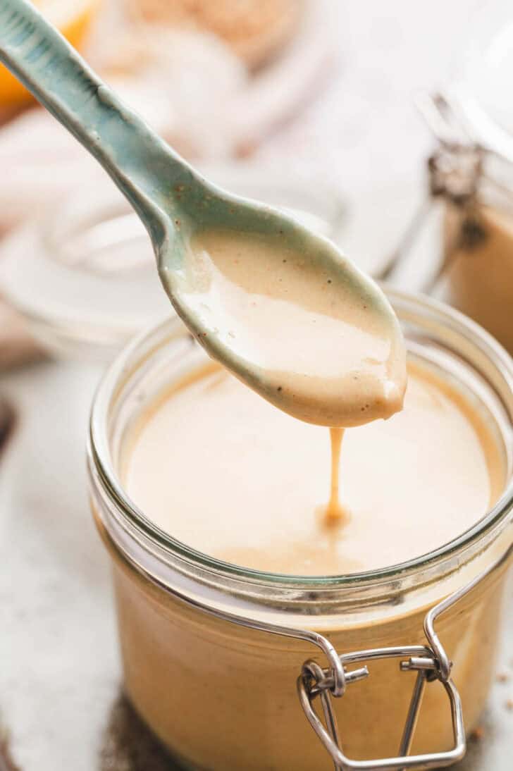 A spoon lifting tahini lemon dressing from a glass jar.