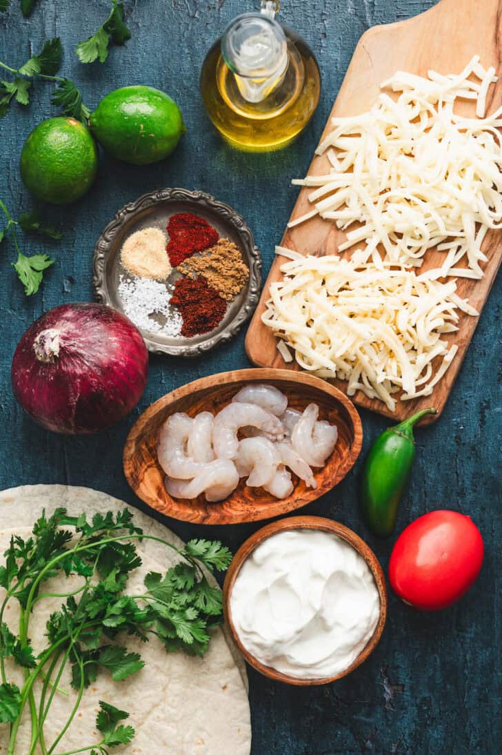 Ingredients on a blue surface, including raw shrimp, shredded cheese, sour cream, vegetables, spices and herbs.