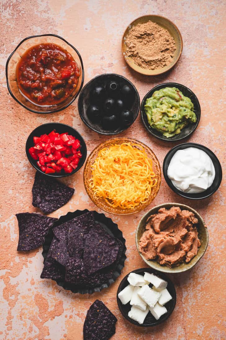Ingredients on a light orange surface, including cheese, beans, cheese, vegetables, guacamole, chips and salsa.