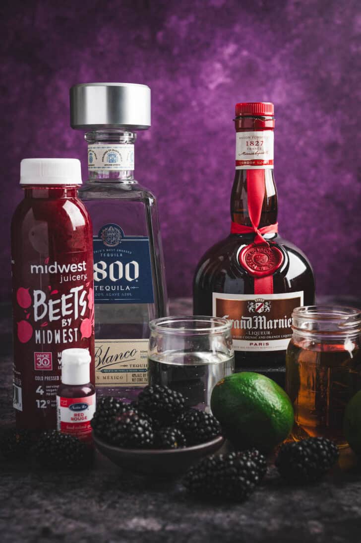 Ingredients in front of a purple background, including tequila, Grand Marnier, beet juice, limes and blackberries.