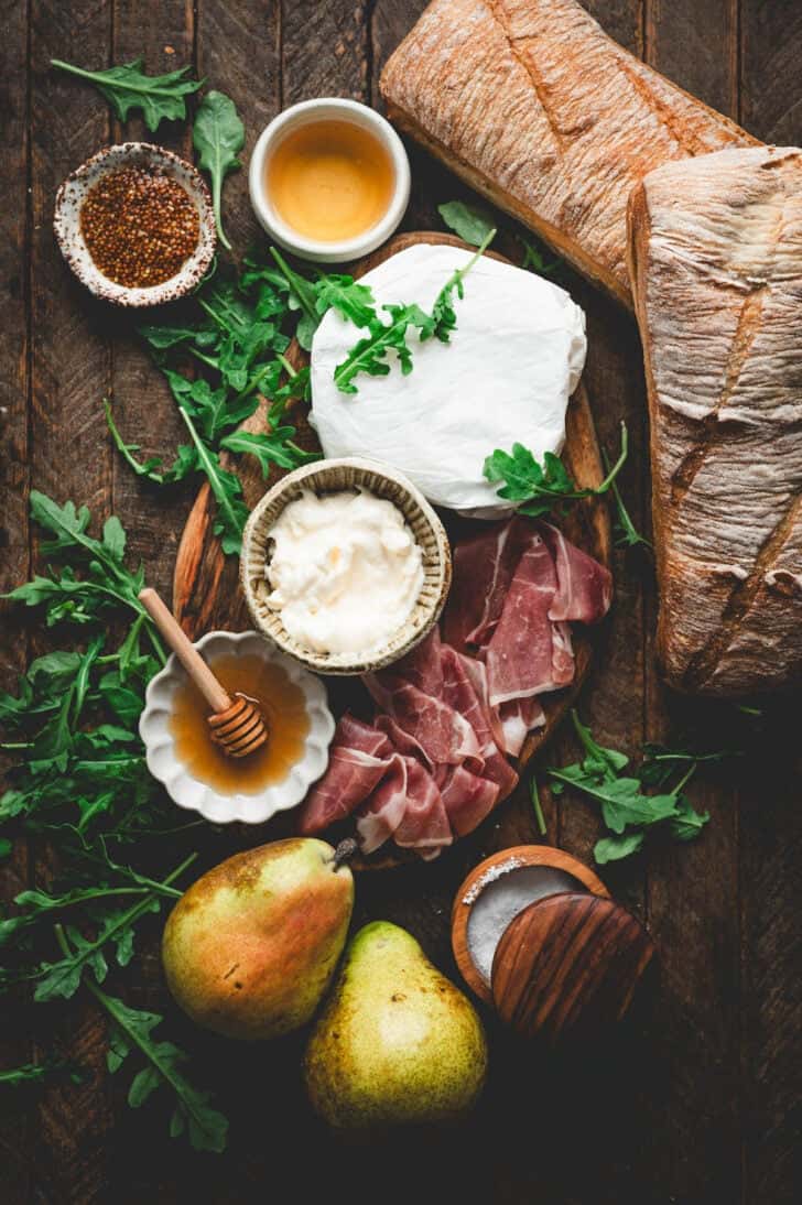 Ingredients on a wooden surface, including charcuterie meat, cheese, honey, pears and arugula.