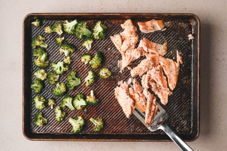 A sheet pan filled with roasted broccoli florets and salmon being broken up with a fork.