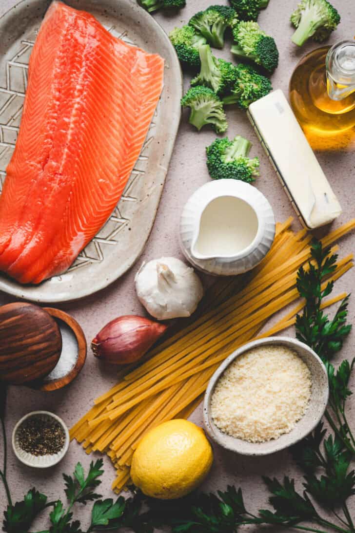 Ingredients on a light background including pink fish, broccoli, pasta, lemon, shallot, garlic, cheese, butter and herbs.