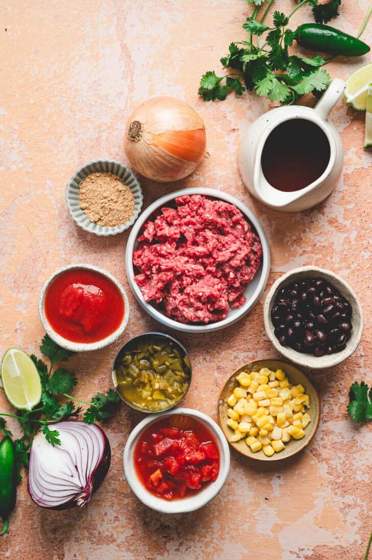 Ingredients on a rustic orange hued background, including ground beef, tomatoes, herbs, corn, onions, peppers and spices.