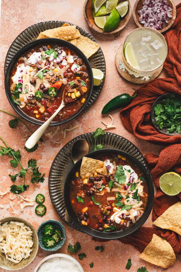 Two bowls of slow cooker taco soup surrounded by toppings for them.