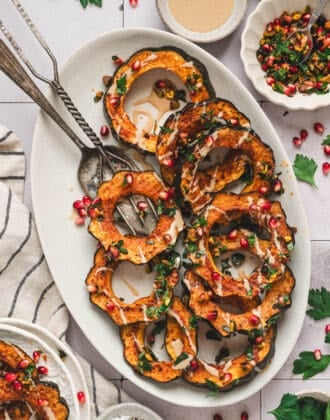 A white oval platter topped with acorn squash roasted and sliced with a creamy drizzle and pistachios and pomegranate seeds sprinkled over the top.