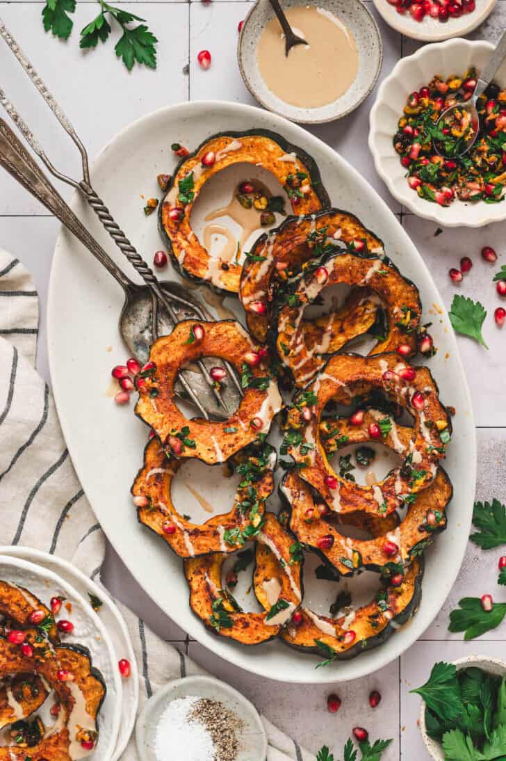 A white oval platter topped with acorn squash roasted and sliced with a creamy drizzle and pistachios and pomegranate seeds sprinkled over the top.