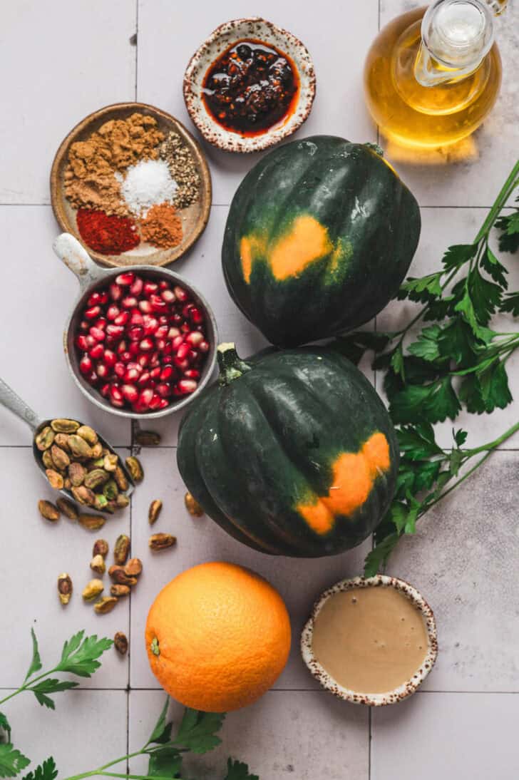 Ingredients on a light tiled background, including fall gourds, orange, pomegranate seeds, oil, herbs and spices.