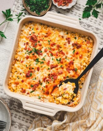 A square baking dish filled with cheesy corn casserole, with a black spoon in it.