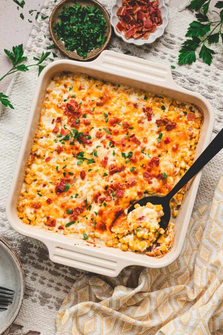 A square baking dish filled with cheesy corn casserole, with a black spoon in it.