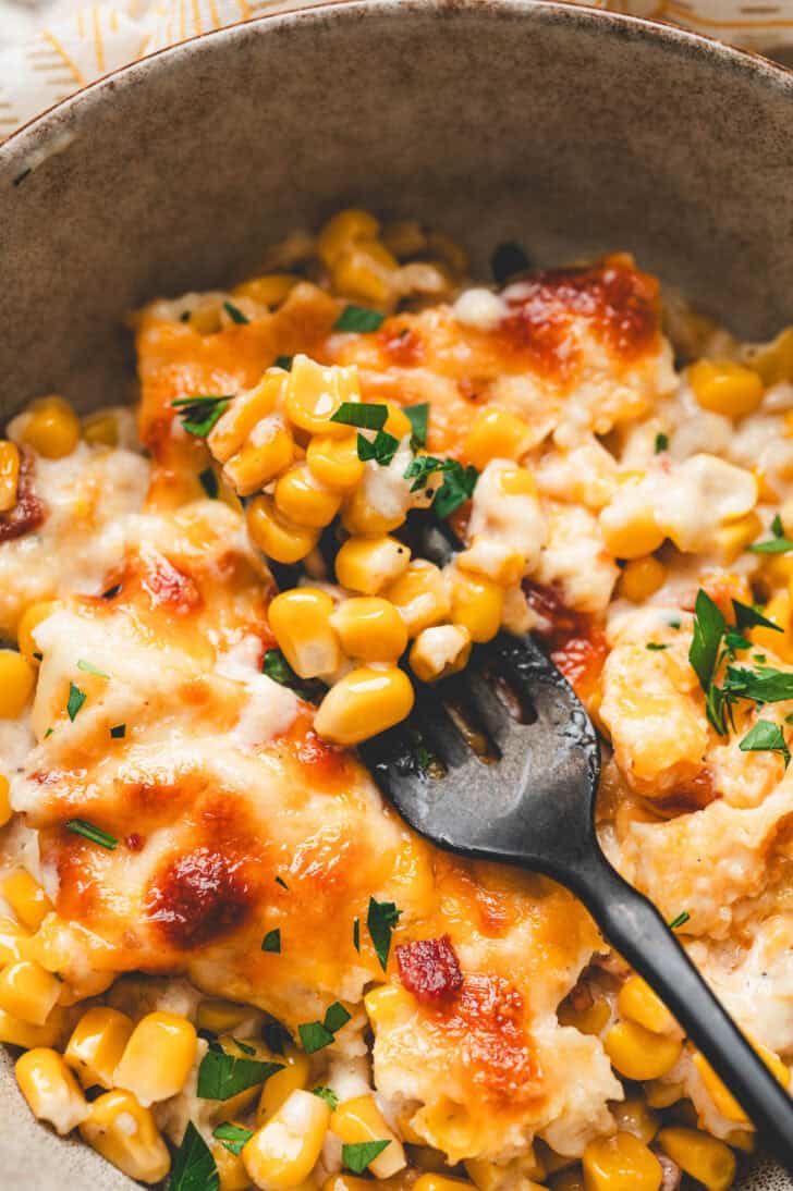 A black fork lifting a bite of corn and cheese casserole from a bowl.