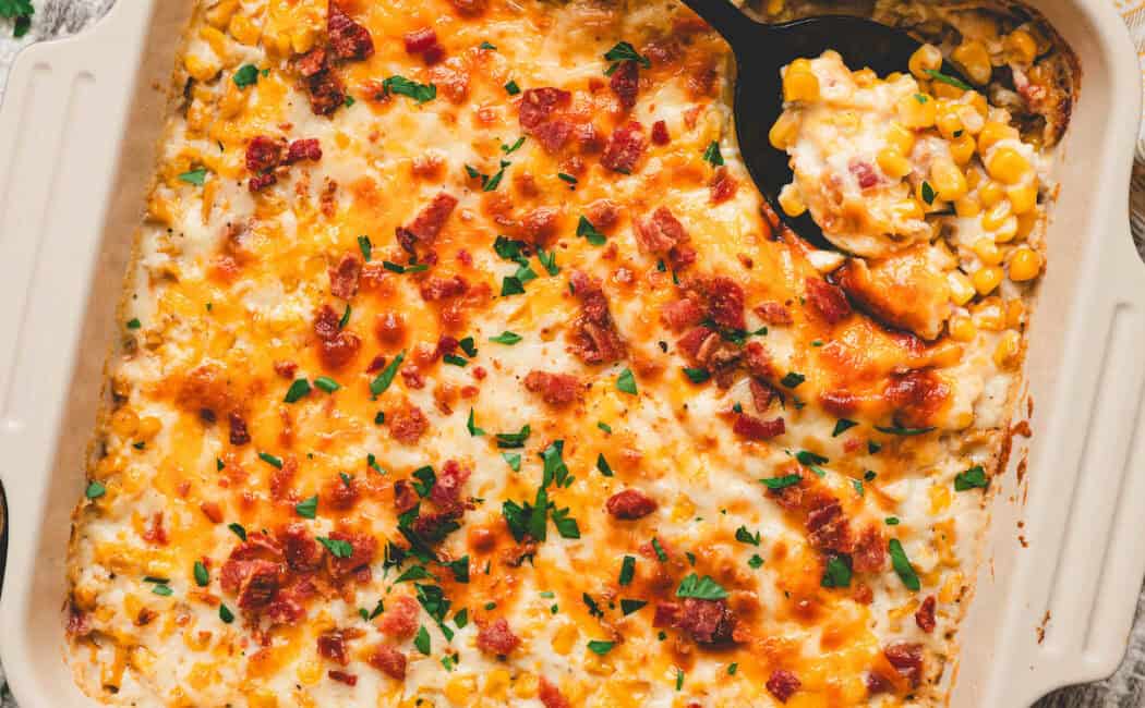A square baking dish filled with cheesy corn casserole, with a black spoon in it.