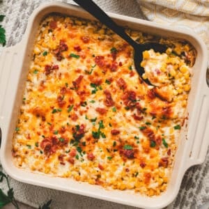 A square baking dish filled with cheesy corn casserole, with a black spoon in it.
