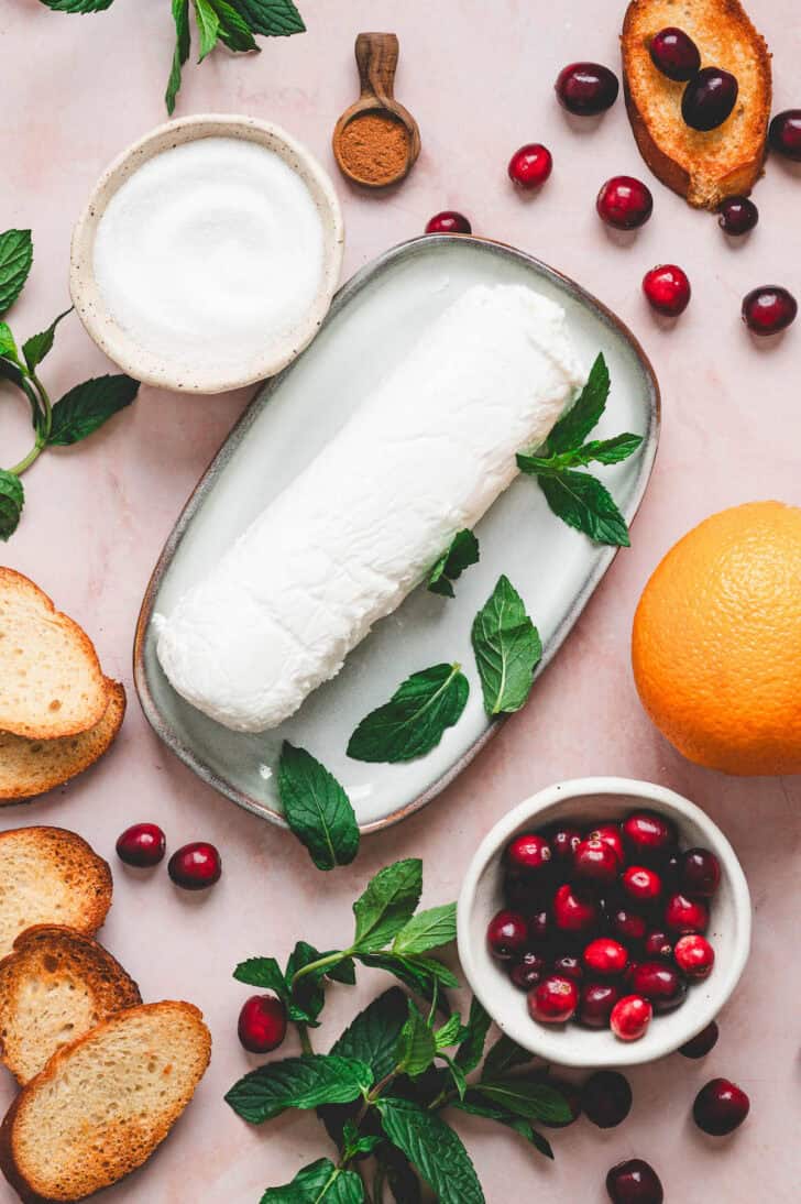 Ingredients for a cranberry appetizer recipe on a light pink surface, including goat cheese, toasted bread, orange and mint.