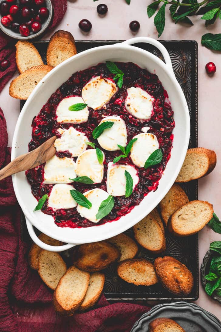A baking dish filled with a goat cheese appetizer with cranberries, surrounded by crostini.