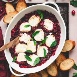 A baking dish filled with a goat cheese appetizer with cranberries, surrounded by crostini.