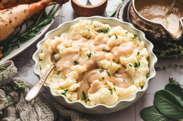 A scalloped edged white bowl filled with mashed potatoes topped with a homemade gravy recipe.